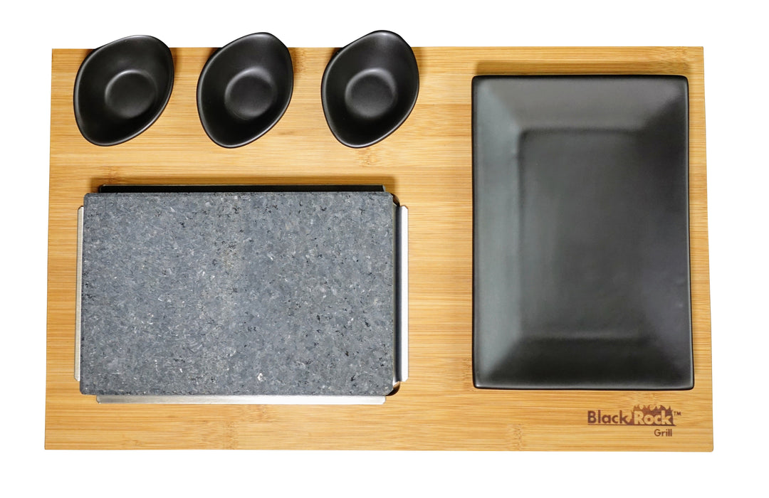 A bamboo serving board with a rectangular stone grill, a large black plate, and three small black dipping bowls arranged neatly on top. The Black Rock Grill logo is in the bottom right corner.