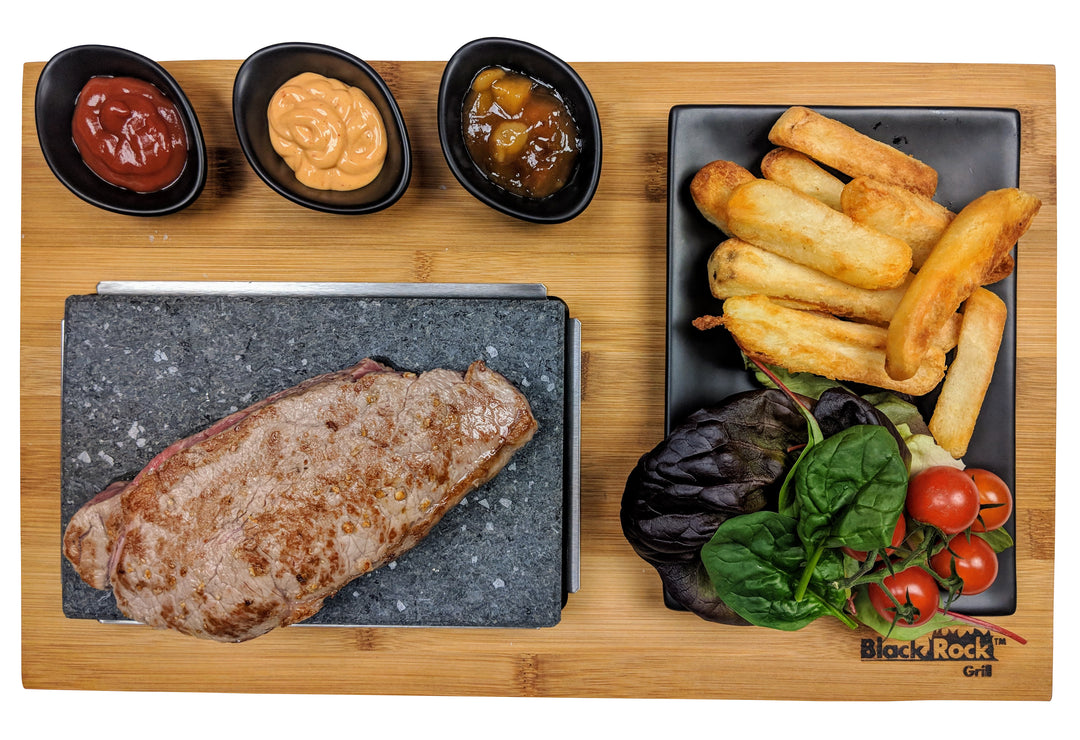 Steak on a hot stone, served with ketchup, sauce, chutney, thick-cut fries, and a salad of lettuce and cherry tomatoes on a wooden board. Black Rock Grill logo visible.