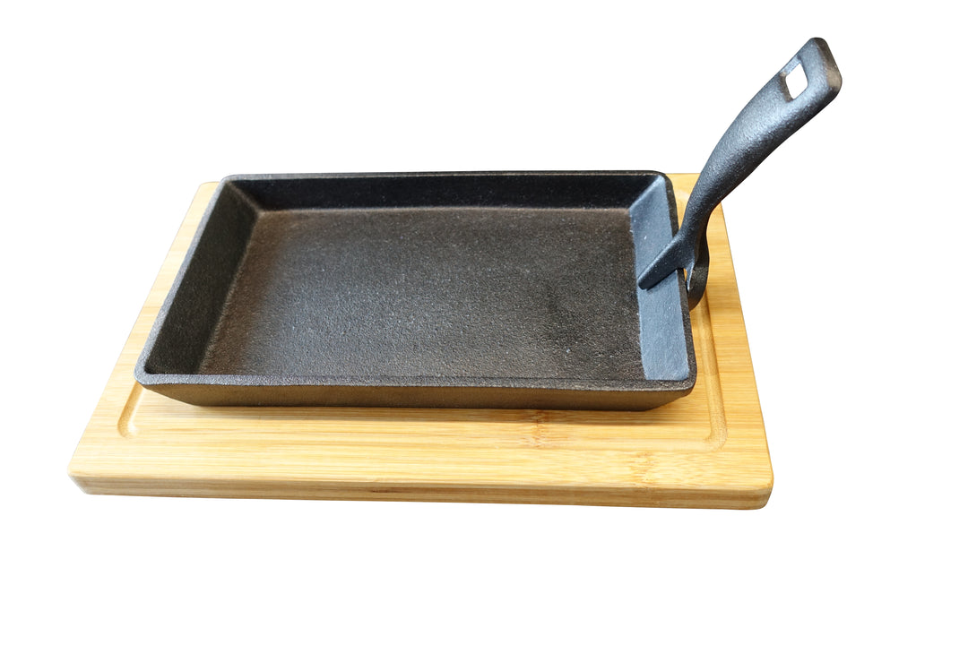 A rectangular cast iron pan with a textured surface rests on a wooden board. The pan has a handle on the right side, angled slightly upwards. The wooden board has a smooth finish.