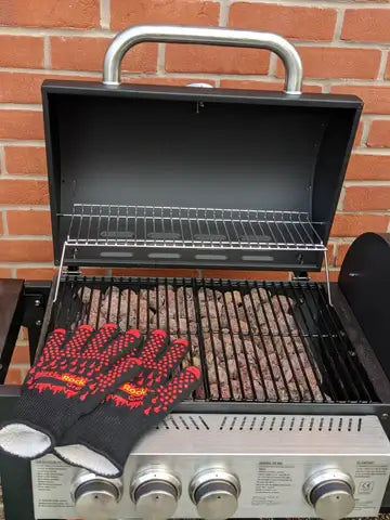 A Black Rock Grill barbecue displays grilled food over Gas BBQ Lava Rocks (4-8cm) with a pair of black and red heat-resistant gloves on the open lid, set against a brick wall.