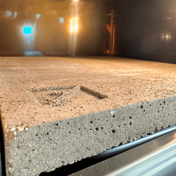 A textured rectangular Effeuno Biscotto Clay Pizza Stone inside an oven, lit by an interior light. It has a small triangular symbol on its side and exudes an industrial feel. Ideal for crafting the perfect Neapolitan pizza.