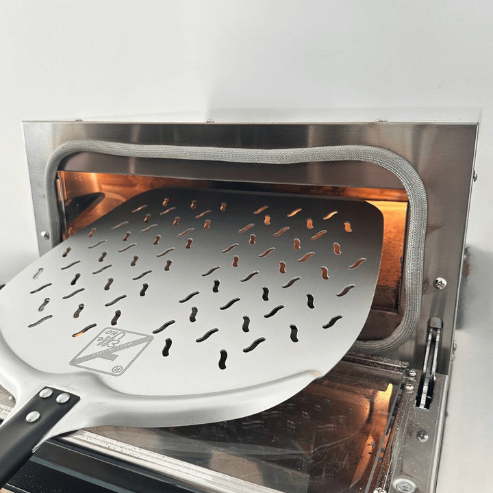 The Effeuno Professional Peel, an anodized aluminum metal pizza peel with a patterned surface, is partially inserted into an illuminated Effeuno Pizza Oven, revealing its sleek silver interior against a white backdrop, perfect for homemade pizza lovers.