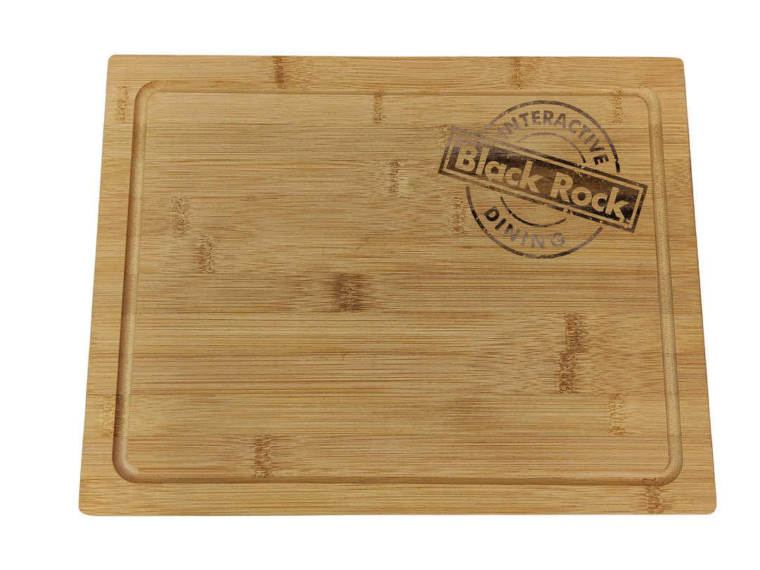 A rectangular bamboo cutting board with a groove, sized 30x20x1.2cm, featuring custom laser engraving of a circular design that reads Black Rock Interactive Dining, by Black Rock Grill.