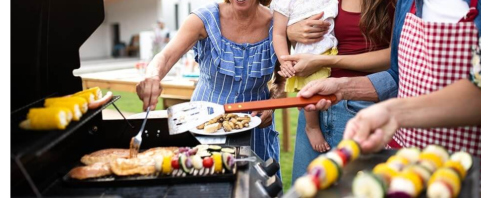 People are gathered around a grill outdoors. One person is turning meat, while another holds a plate of vegetables and corn. A baby is held by someone wearing a blue and white dress. Bright and sunny setting.