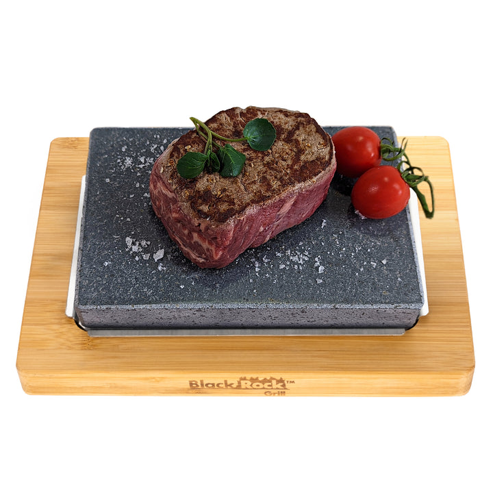 A seasoned steak sizzles on a hot stone slab, garnished with herbs, alongside two vine-ripe tomatoes for color. This tantalizing setup on the wooden board proudly displays Black Rock Grill, ideal for those who cherish Steak Stone Cooking with the Steak Stone Cooking Set from Black Rock Grill.