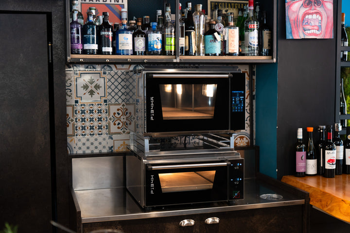 Two black Effeuno ovens with the Black Rock Grill Stacking Kit for PRO & Evolution Ovens are double-mounted on a stainless steel counter under a shelf with bottles of alcohol, in a bar or restaurant with a patterned tile backsplash.