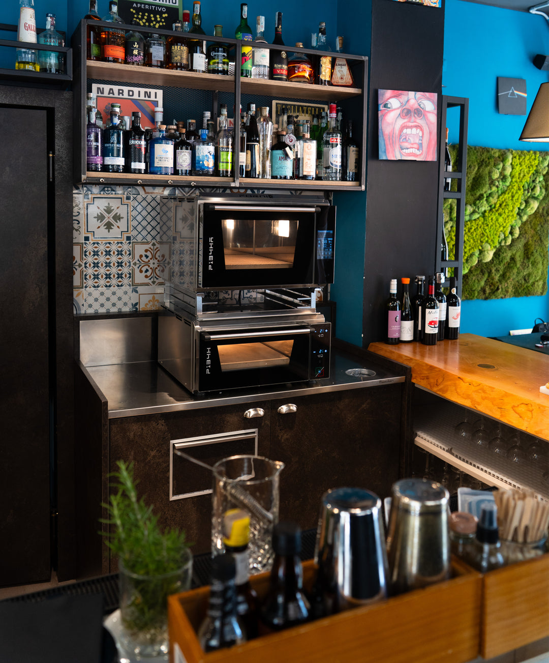 A bar area with shelves of liquor bottles, glassware, cocktail shakers, and wine bottles features a Black Rock Grill Effeuno Stacking Kit for PRO & Evolution Ovens as a safe double oven mount, plus a wall with green moss and patterned tiles.