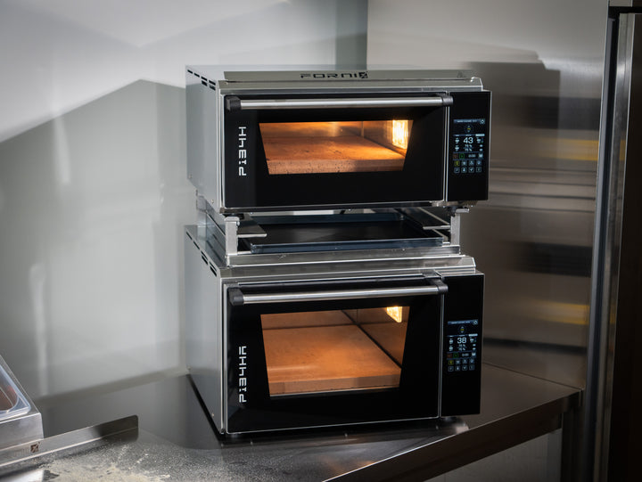Two Effeuno PRO & Evolution ovens are stacked on a stainless steel table using the Black Rock Grill Stacking Kit for a safe double oven mount. Their digital panels are lit, showing empty stone baking surfaces inside.