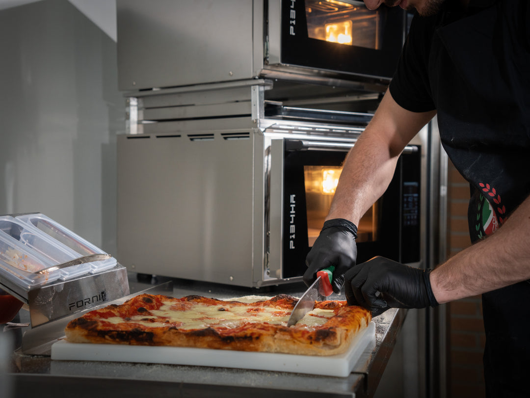 Wearing black gloves, a person slices a rectangular pizza on a white cutting board in a kitchen with the Black Rock Grill Effeuno Stacking Kit for PRO & Evolution Ovens – Safe Double Oven Mount in the background.
