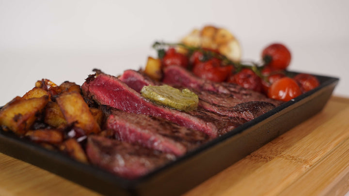 A close-up of medium-rare steak with herb butter on a Cast Iron Fajita Skillet Pan by Black Rock Grill, featuring roasted cherry tomatoes, garlic, and potato wedges. Served on its wood base, this premium dish is displayed on a wooden surface for an elegant presentation.