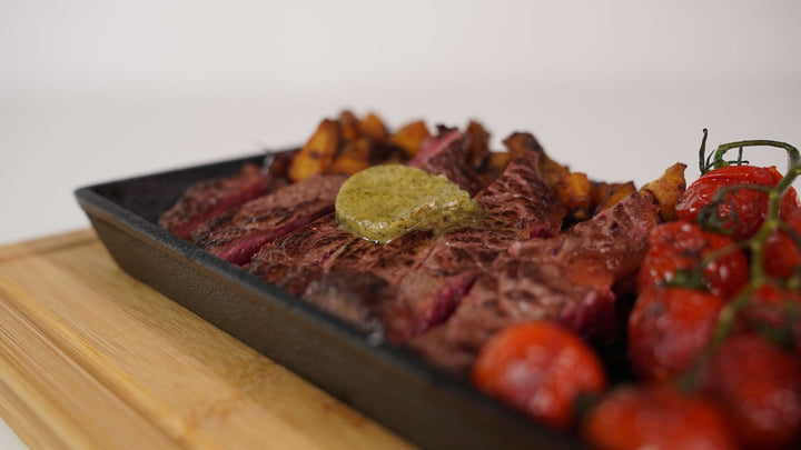 Close-up of a premium steak topped with herb butter on a Black Rock Grill Cast Iron Fajita Skillet Pan, served with roasted potatoes and vine-ripened cherry tomatoes on its wood base.