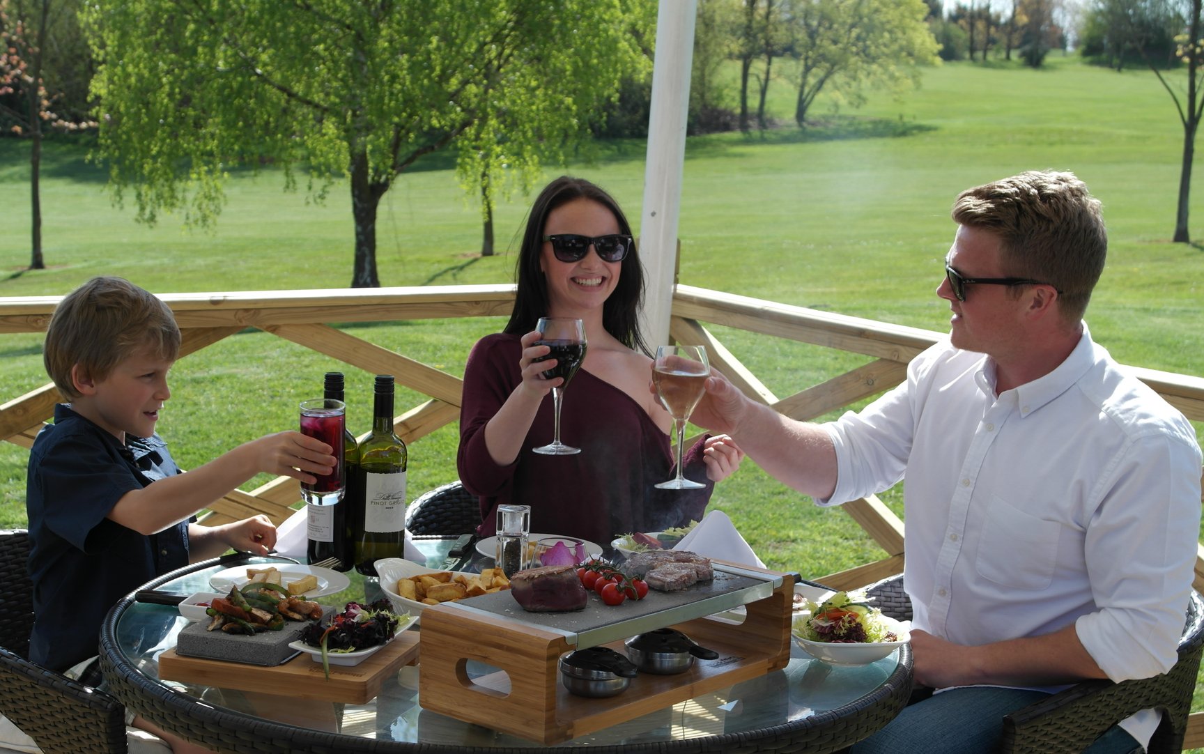 A family of three enjoys an outdoor meal on a sunny day. They sit at a round table with wine, steak, and various foods, toasting with drinks. A green lawn and trees are in the background. The atmosphere is relaxed and cheerful.