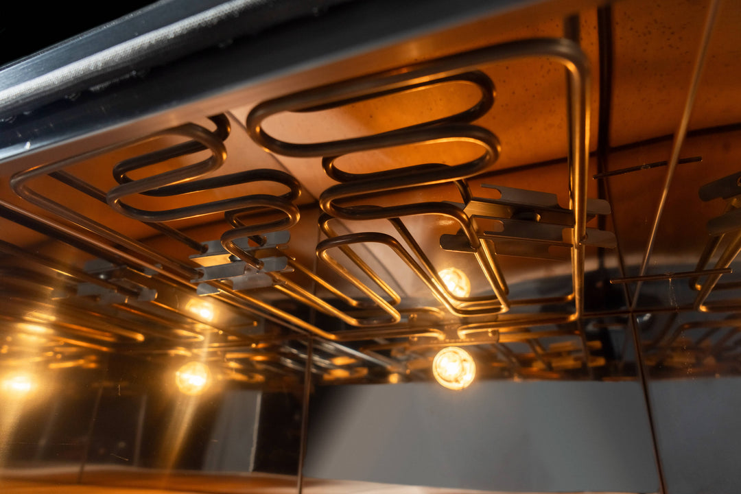 A close-up reveals the interior of the Effeuno Pizza Oven P134H 509 PRO + Biscotto Stone, where glowing heating elements form intricate patterns. The warm light reflects off shiny surfaces, showcasing craftsmanship typical of professional pizza ovens.