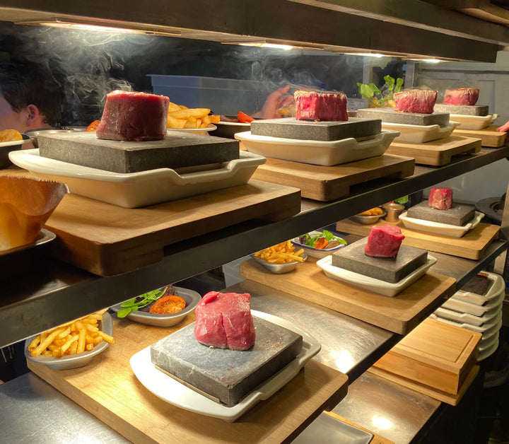 The Black Rock Grill’s ROXY12 set showcases raw meat on hot stone slabs in white dishes atop wooden boards, providing an interactive experience with steaming stones. Sides like fries sit nearby as a person partially visible cooks at the table in the kitchen.
