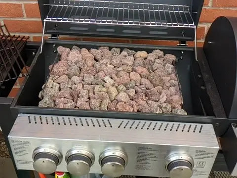 The Black Rock Grill Lava Rocks for Grilling (4–8cm) are shown inside a gas grill with four control knobs and an open lid, creating an ideal cooking surface. A warming rack sits above the rocks, with a brick wall as the backdrop.