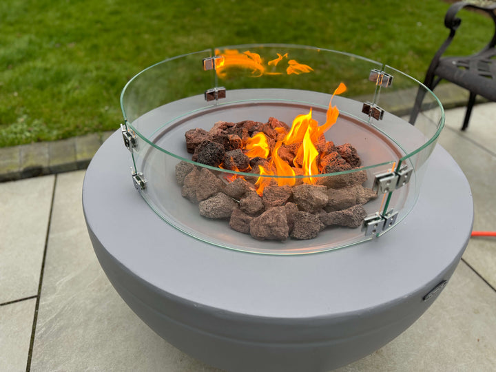 A modern gas fire pit with bright yellow-orange flames and Black Rock Grill's 100% natural Lava Rocks (4-8cm) sits on a stone patio, encircled by a circular glass wind guard, with green grass in the background.
