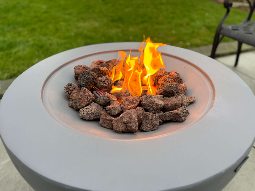 A close-up of Black Rock Grill's 100% Natural Lava Rocks for Firepit (4-8cm) with bright flames among the rocks, set on a patio with green grass and part of a metal chair visible in the background.
