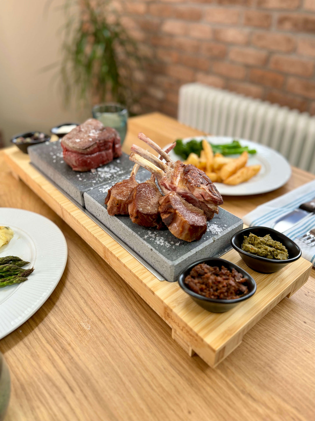 Signature Steak Sharing Stone Set with bamboo board, 2 Steak stones and 4 dipping ramekins. Steak is cooking on the stones and set is put on a dining table ready for a meal