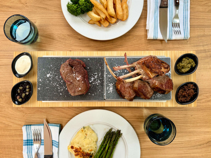An overhead view of a dining table showcases the Black Rock Grills Signature Steak Sharing Stone Set with steak, lamb chops, fries, broccoli, and sauces. Visible are plates of asparagus and gratin with cutlery alongside two glasses of water.