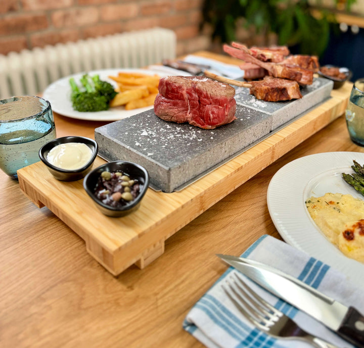 Signature Steak Sharing Stone Set with bamboo board, 2 Steak stones and 4 dipping ramekins. Steak is cooking and the set is put on a dinning table ready for a meal