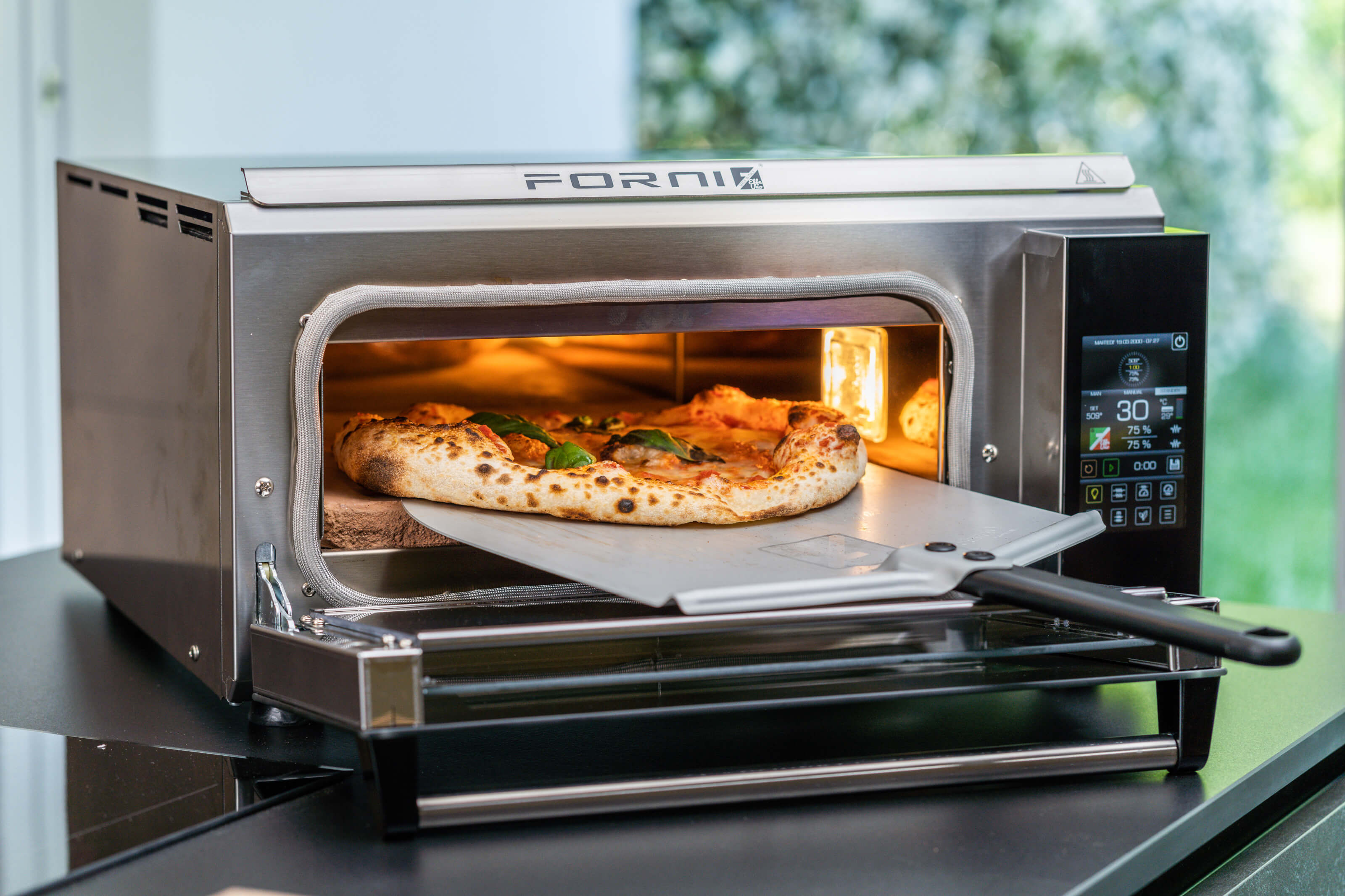 A pizza being baked in a modern stainless steel oven with digital controls. A pizza peel with a wooden handle is partially inserted, holding the pizza inside the oven. The pizza is topped with basil and cheese.