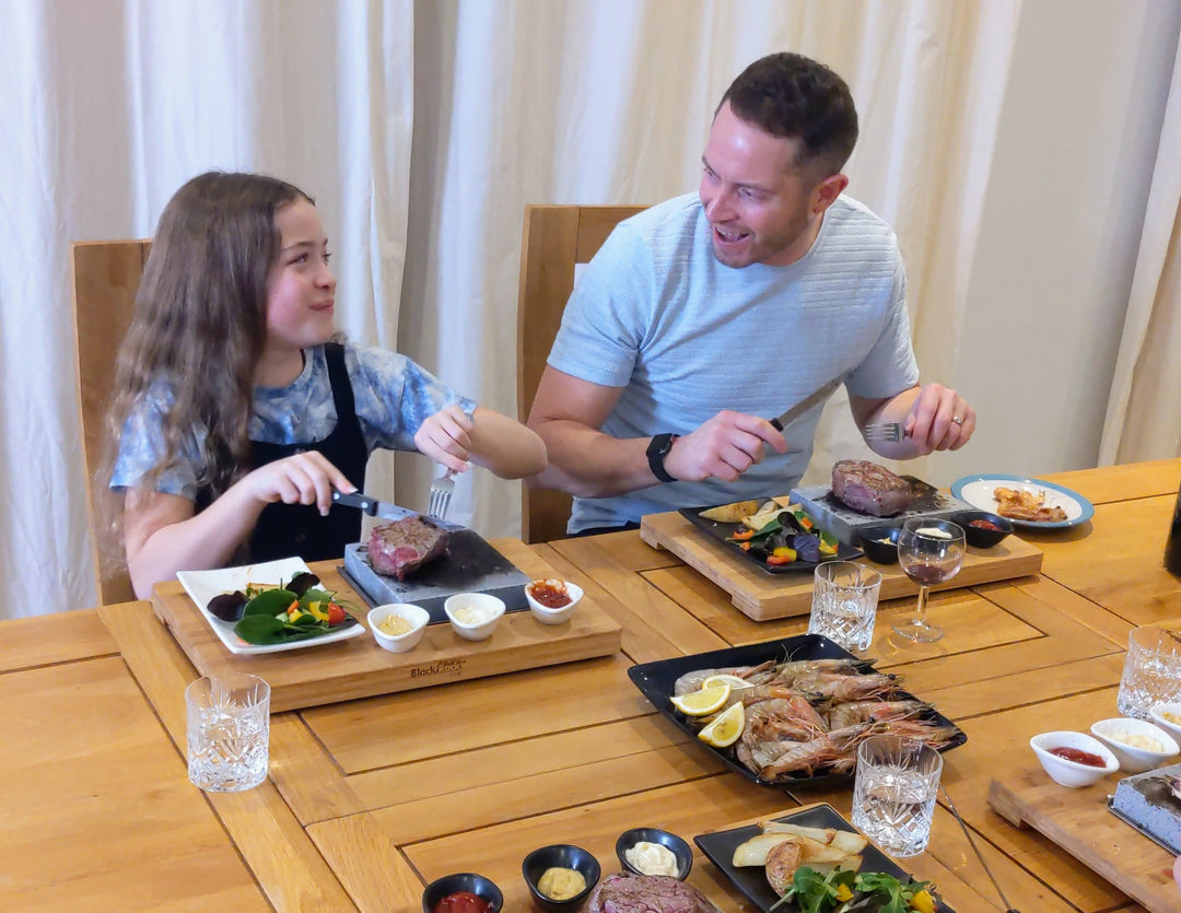 A man and a girl are sitting at a dining table, smiling at each other. They have plates of steak and vegetables. The table is set with dishes of prawns, condiments, and glasses of water. White curtains are in the background.