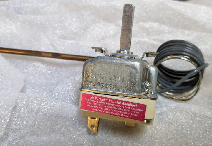 The Effeuno 450°C Thermostat by Black Rock Grill, compatible with models P134H, P134HA, P150H, P150HA, and F50, sits on bubble wrap. It has a coil, protruding lever, multilingual caution label, visible numbers, and electrical connectors.
