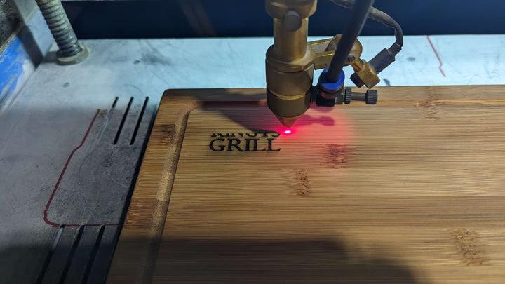 A laser engraving machine etches MINUTE GRILL onto a Black Rock Grill Wooden Serving Steak Board, creating a red dot to show the area being engraved. Ambient industrial lighting illuminates the eco-friendly bamboo.
