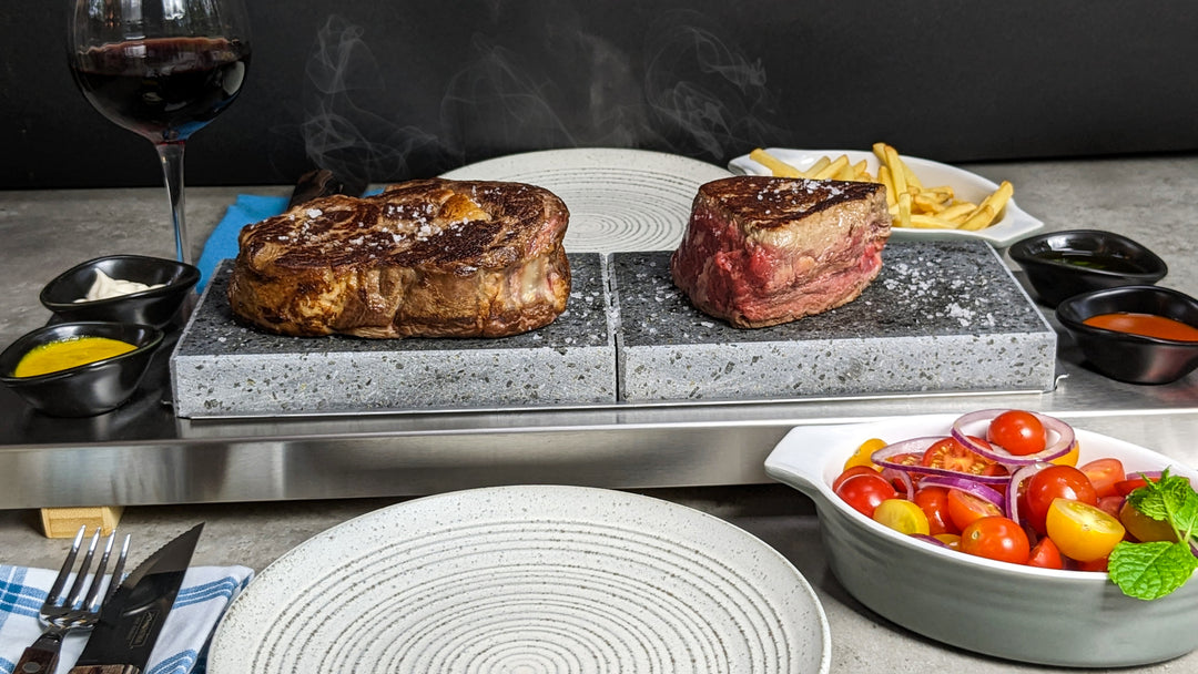 one ribeye and one fillet steak cookig onthe hot rock on the tbale with wine and chips
