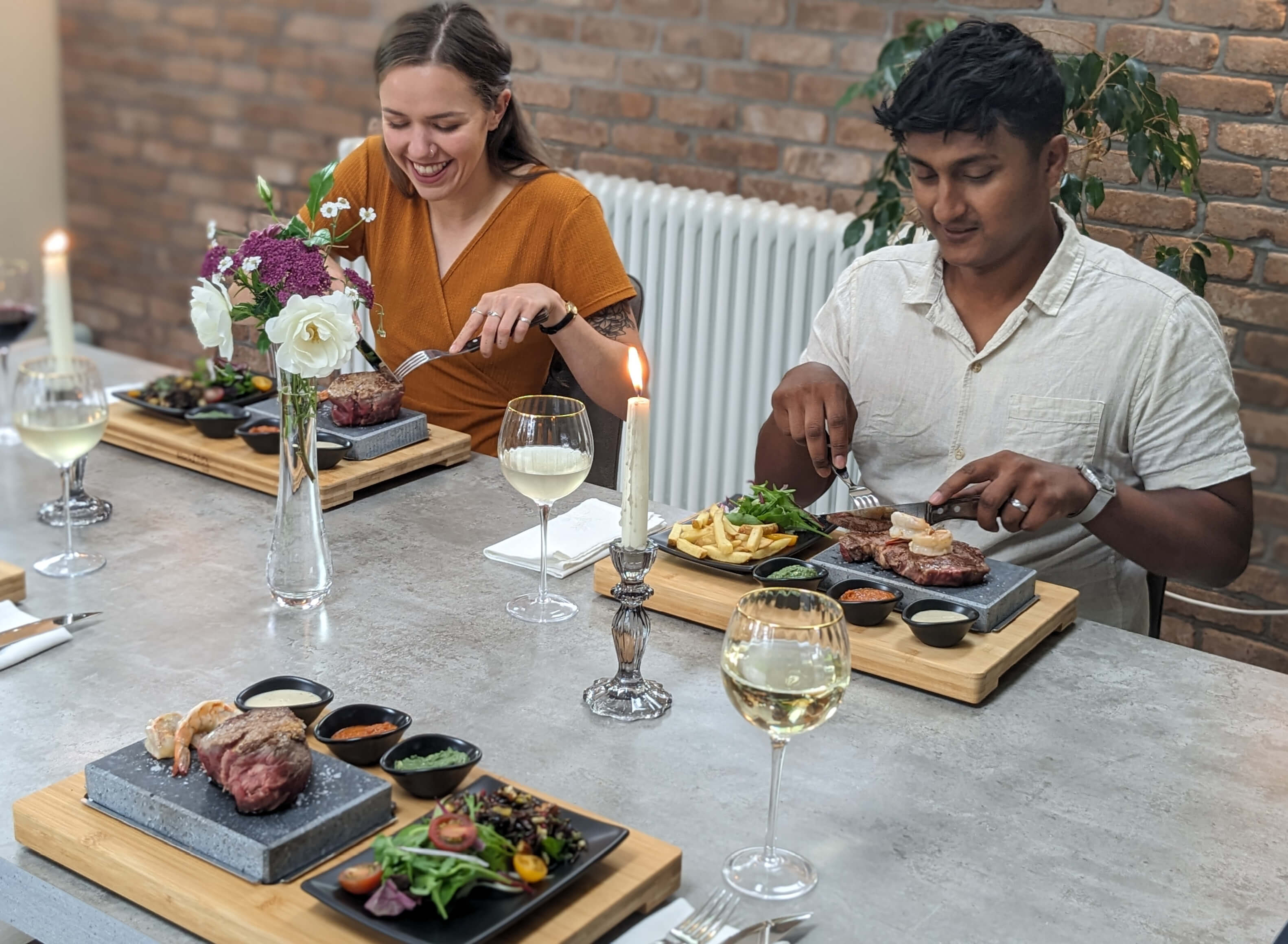 Two people are seated at a dining table, enjoying a meal. Each has a hot stone platter with meat and sides. There are flowers and candles on the table, and glasses of white wine. They appear to be engaged in conversation and are smiling.