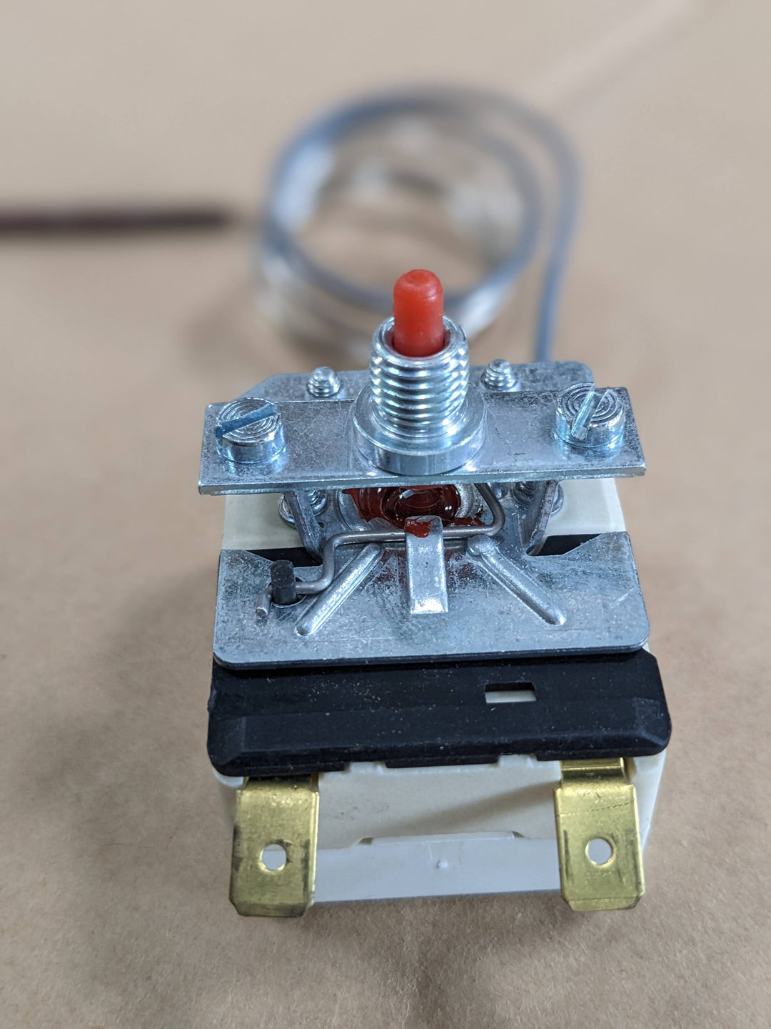 The close-up shows a Black Rock Grill Overheat Reset Switch for Effeuno P134H ovens with a red button and brass terminals. Behind it, the blurred background features a coiled metallic cable, designed to prevent overheating up to 515°C.