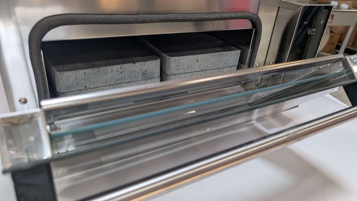 A close-up of the Black Rock Grill Steak Stone Grill & Effeuno Pizza Oven Combo Package shows the open stainless steel oven with firebrick blocks and a Biscotto stone, highlighting the door and interior for authentic hot stone cooking.