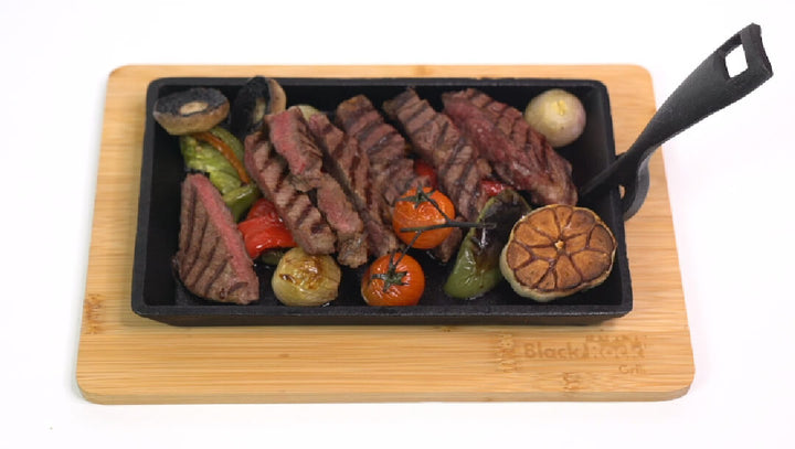 The Black Rock Grill Cast Iron Fajita Skillet Pan with Wood Base includes sliced steak, grilled mushrooms, tomatoes, bell peppers, and roasted garlic served on a wooden board.