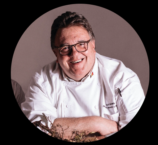 A smiling chef wearing glasses and a white chefs coat stands against a neutral background. The coat has a small flag detail on the collar.