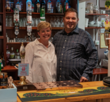 A woman and a man stand smiling behind a bar counter. Various bottles and barware are visible on shelves behind them. The counter is decorated with placemats and a small sign.