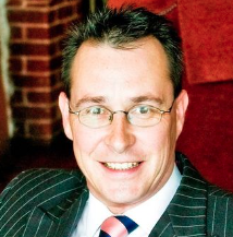 Man in a pinstriped suit and tie, wearing glasses, smiling at the camera. The background features a red and brick wall.