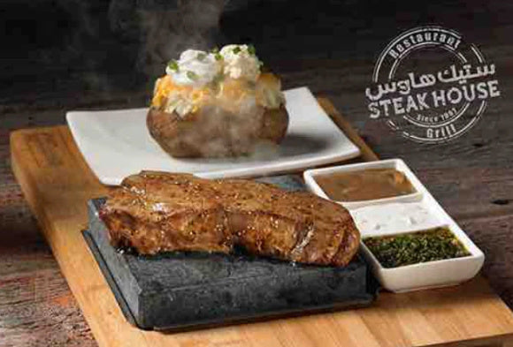 A steaming grilled steak on a hot stone is served on a wooden board. Next to it is a baked potato topped with sour cream and chives on a separate plate. Accompanying sauces are placed beside the steak. Steakhouse grill branding is visible.