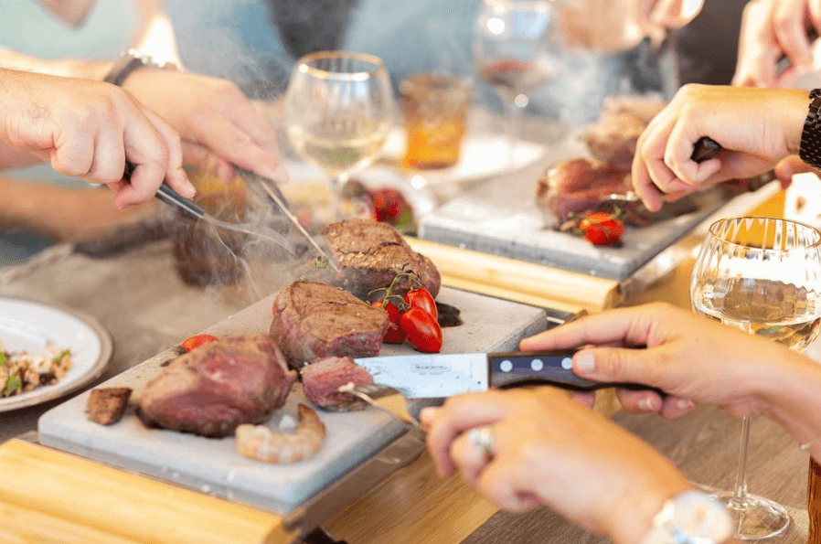 Hands use utensils to cut and serve pieces of steak on hot stones, surrounded by a few vegetables and glasses of wine. The table setting is lively, suggesting a shared dining experience.