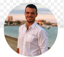 A person with short dark hair and a beard is smiling, wearing a white collared shirt. The background shows a waterfront with boats and a cityscape under a blue and pink sky.