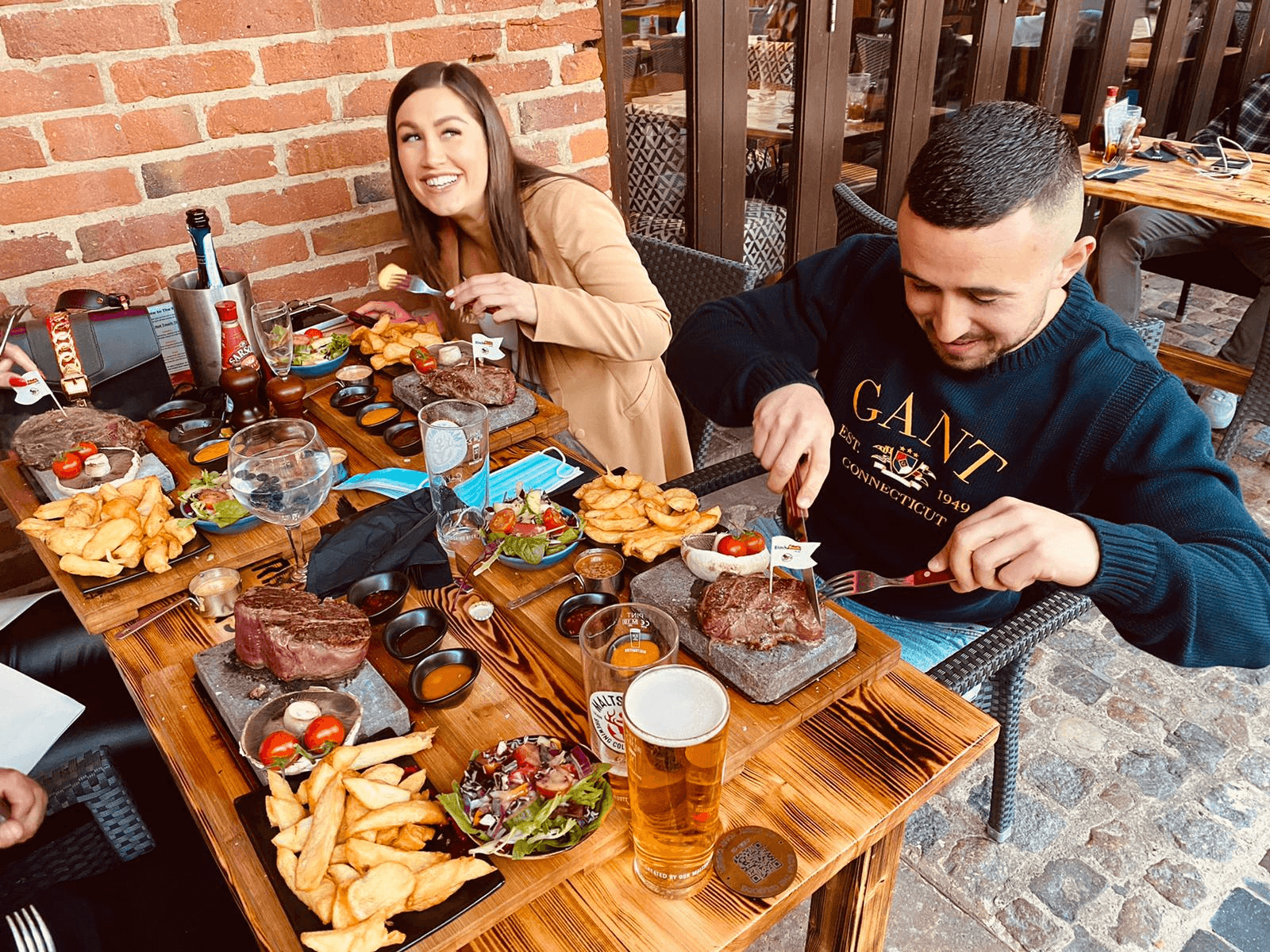 A man and a woman sit at a wooden table, enjoying meals with steak, fries, and other sides. The man is cutting his steak, while the woman is smiling. There are drinks on the table, including a beer and a gin and tonic. The setting is casual and outdoors.
