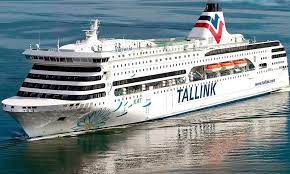 A large white cruise ship with Tallink written on the side sails on calm blue waters. The ship features multiple decks, a blue and red funnel, and lifeboats visible along the side. The sea is smooth, reflecting the ships image.