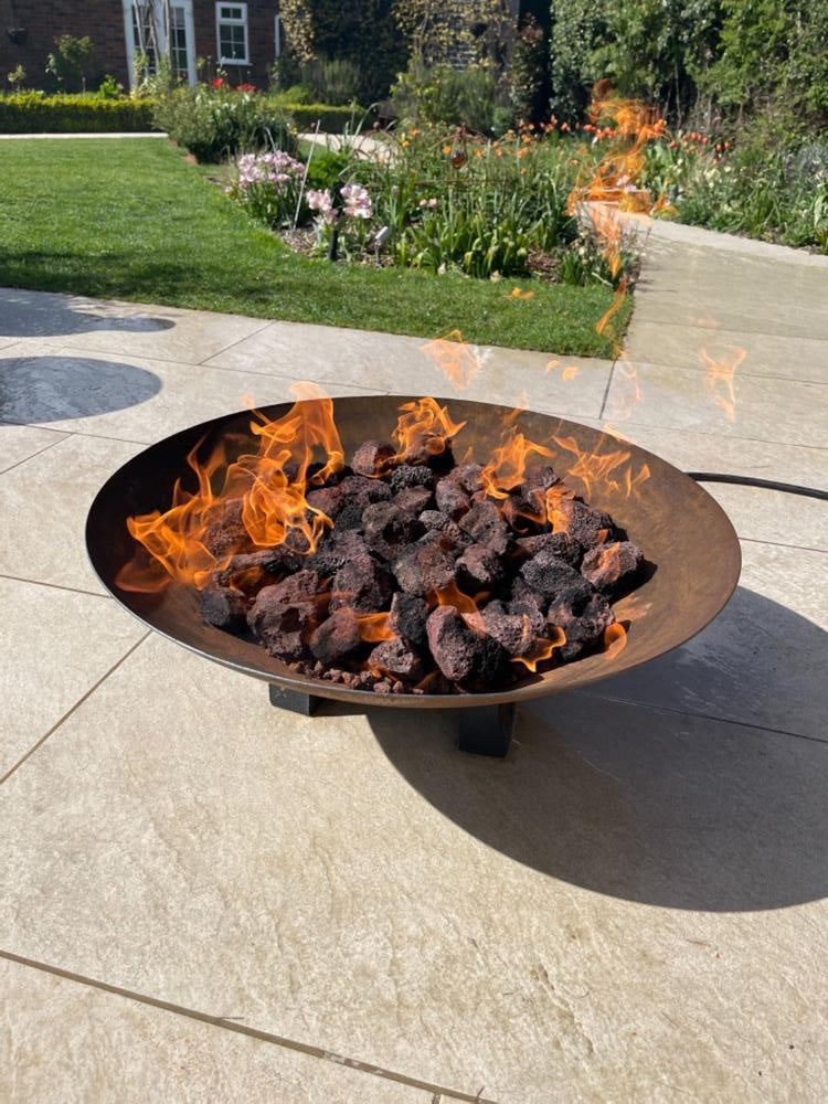 A Black Rock Grill firepit, filled with 100% natural Lava Rocks for Firepit (4-8cm), sits on a stone patio in a sunlit garden, with grass, flowers, and a house in the background.
