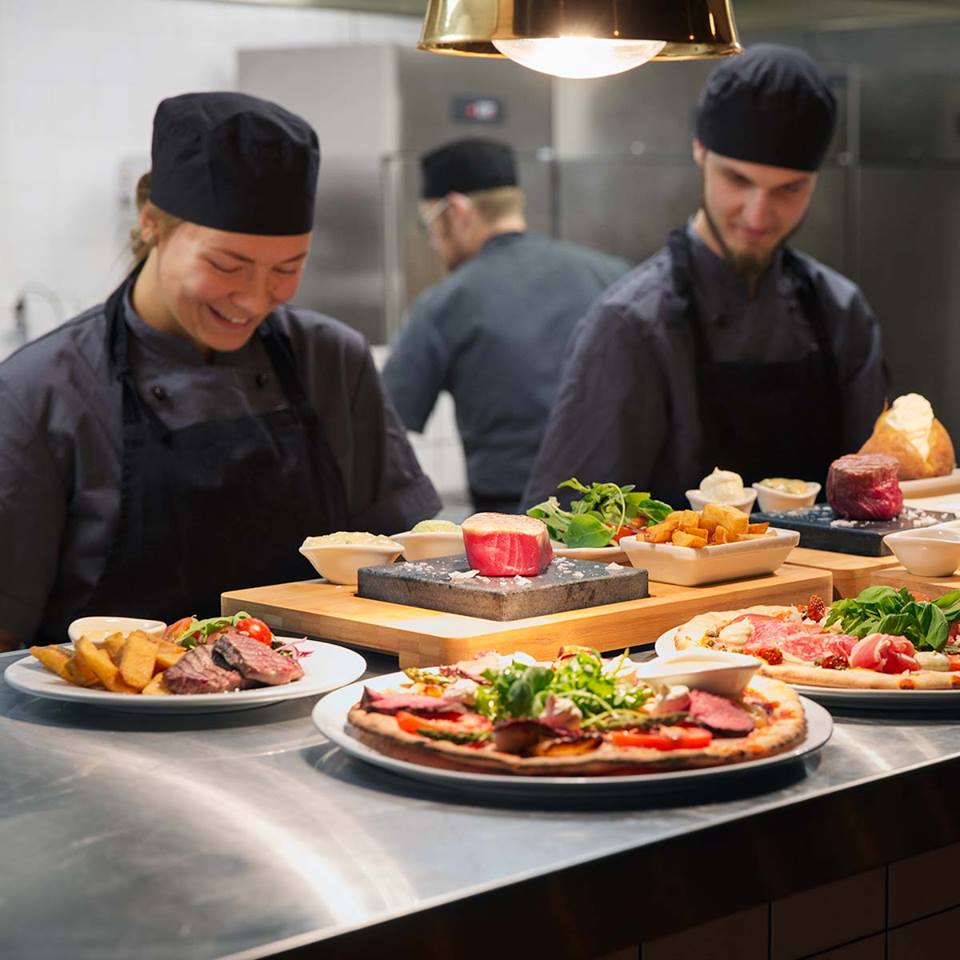 Two chefs in black uniforms and hats prepare food in a commercial kitchen. In the foreground, plates with pizza and steak are displayed. A third chef works in the background near a heated lamp.