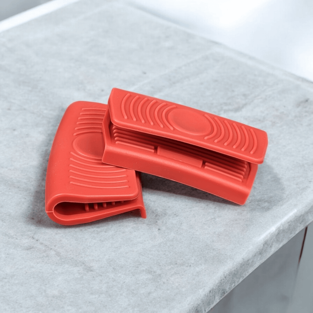 A pair of Black Rock Grill Silicone Heat Resistant Handle Holder Covers, made from premium food-grade silicone with ridged surfaces, rest on a light gray marble countertop.