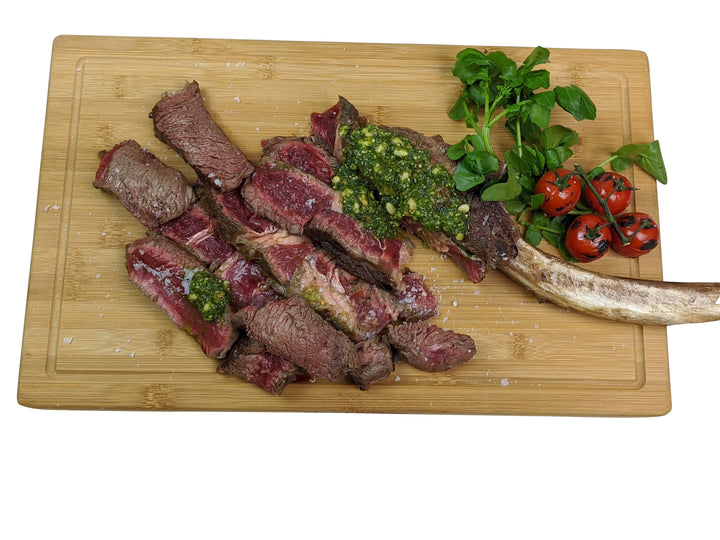 Medium-rare sliced steak with green sauce on a Black Rock Grill large wooden serving board, complemented by a grilled bone and charred cherry tomatoes with greens.