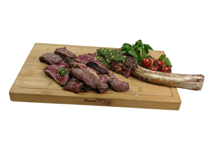 Medium-rare steak with chimichurri sauce is elegantly displayed on a Black Rock Grill Wooden Serving Board. A large bone and vine tomatoes enhance the dish, while fresh greens complete the presentation.