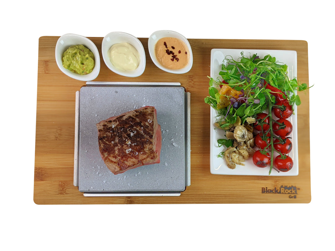 The Black Rock Grill Steak On The Stone Set invites hot stone dining enthusiasts to cook their meal on a wooden board with a sizzling steak and three sauces (avocado, mayo, and spicy orange). A fresh salad with tomatoes, greens, and mushrooms adds vibrant colors to the side.