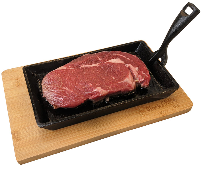 A raw slice of beef sizzles on the Black Rock Grill Cast Iron Fajita Skillet Pan with its black rectangular surface elegantly resting on a wooden base.