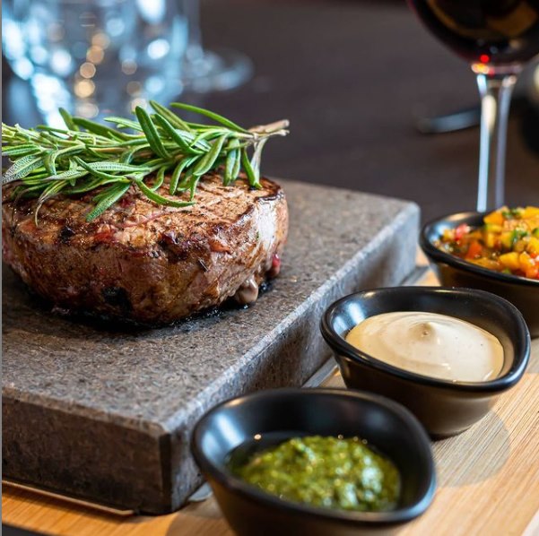 A ROXY45 from Black Rock Grill showcases live cooking artfully with a juicy steak on a stone slab. Three bowls offer green and white sauces plus diced veggies for a healthy meal. A glass of red wine in the background elevates this interactive experience with the 45 Rock, 45 Plate, and Rock Oven set up.