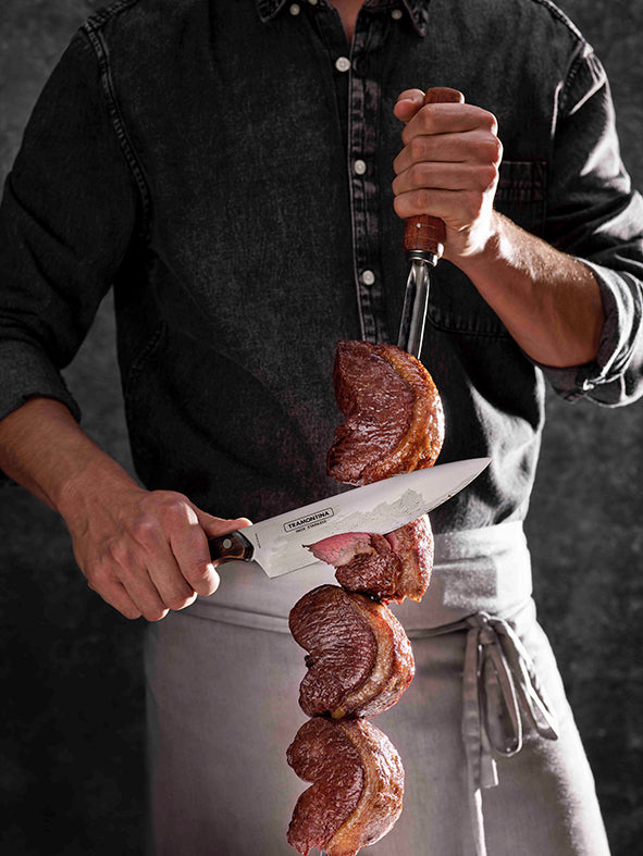 Wearing a dark shirt and apron, a person deftly slices juicy, well-cooked meat from a Tramontina Churrasco BBQ Skewer 750mm using a large stainless steel knife, crafting an appetizing and rustic culinary scene.